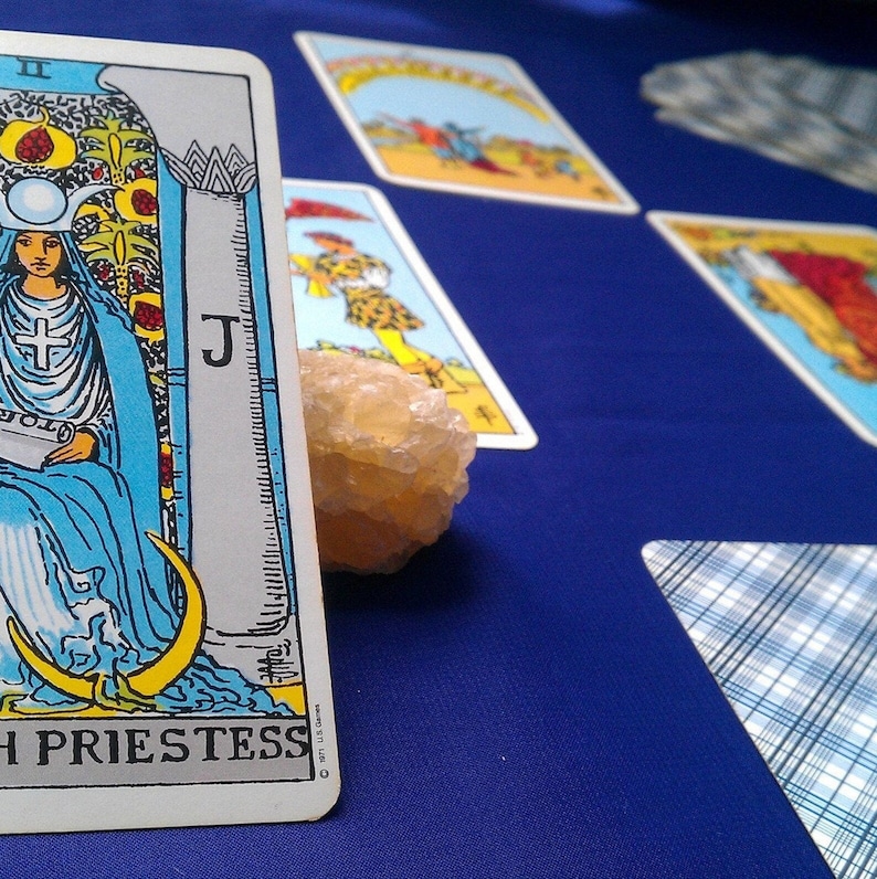 May include: The High Priestess tarot card features a woman in a blue robe sitting on a throne with a crescent moon on each side. The card is part of a tarot card spread on a blue surface.