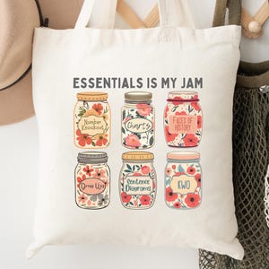 May include: A cream-colored tote bag with the text "ESSENTIALS IS MY JAM" and illustrations of six mason jars filled with flowers. A tan hat and two other bags hang on a wooden rack. The Polka Dottie Studio logo is in the lower left corner.