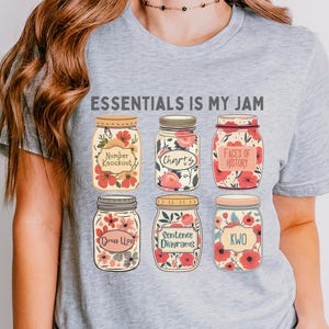 May include: Athletic heather gray t-shirt with the text "ESSENTIALS IS MY JAM." The shirt features a graphic of six mason jars with floral designs and labels like "Number Knockout" and "Charts." The Polka Dottie Studio logo is in the upper right corner.