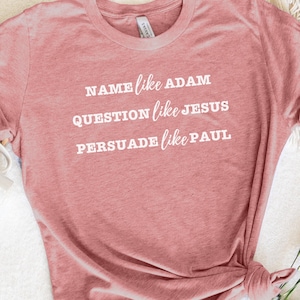 May include: Heather mauve t-shirt with the text "NAME like ADAM, QUESTION like JESUS, PERSUADE like PAUL" printed in white.
