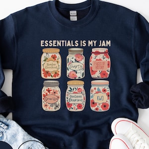 May include: Navy blue sweatshirt with the text "ESSENTIALS IS MY JAM." The design features six mason jars filled with flowers and labels like "Number Knockout" and "Charts." A Polka Dottie Studio logo is visible.