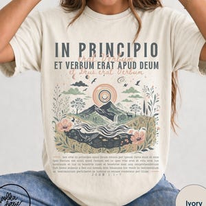 May include: Ivory-colored t-shirt featuring a landscape design with mountains, a rainbow, and floral elements. The shirt has the text "IN PRINCIPIO" and other Latin text. The shirt is from Polka Dottie Studio.