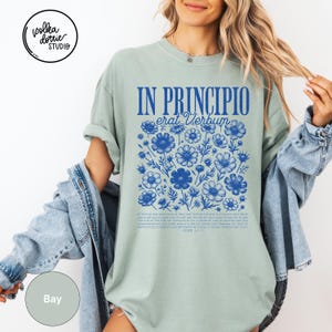 May include: A light green t-shirt with the text "IN PRINCIPIO erat Verbum" in blue, along with a floral design. The shirt also features additional text at the bottom. The Polka Dottie Studio logo is visible in the upper left corner.