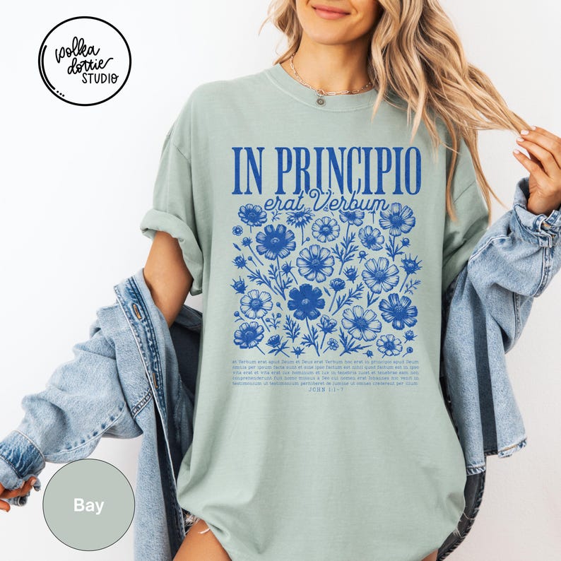 May include: A light green t-shirt with the text "IN PRINCIPIO erat Verbum" in blue, along with a floral design. The shirt also features additional text at the bottom. The Polka Dottie Studio logo is visible in the upper left corner.