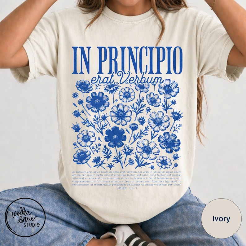 May include: Ivory-colored t-shirt with a blue floral design and the text "IN PRINCIPIO erat Verbum". The design includes a cluster of blue flowers and Latin text. The shirt is from Polka Dottie Studio.