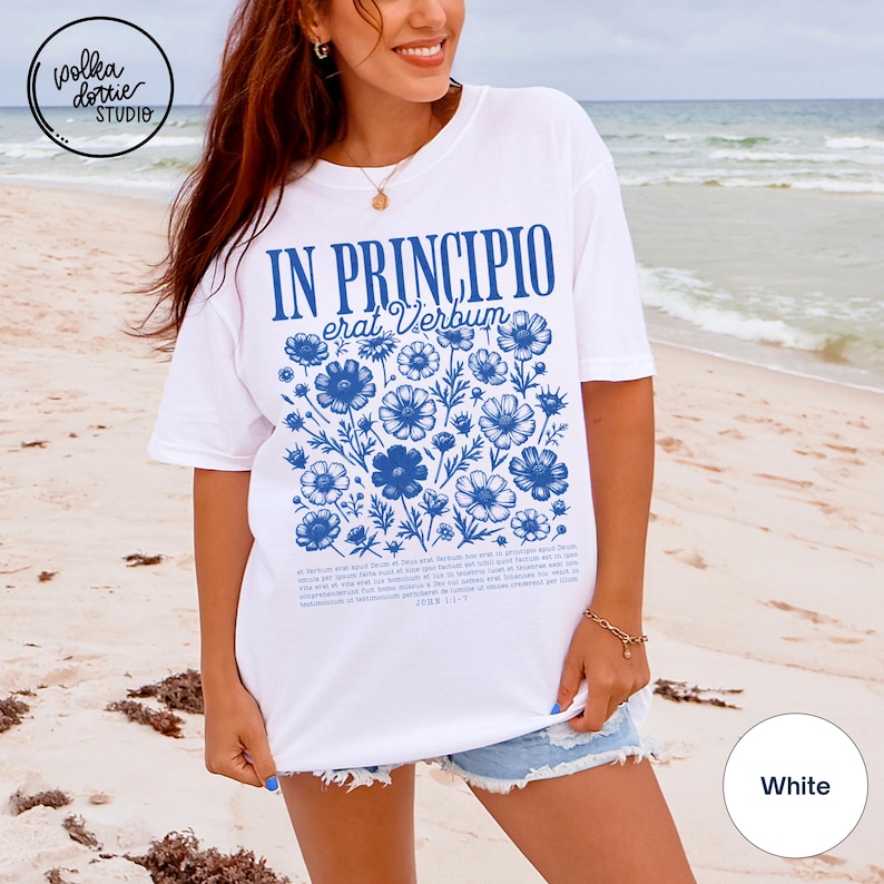 May include: White t-shirt with a blue floral design and the text "IN PRINCIPIO erat Verbum". The design includes blue flowers and text from John 1:1-7. The shirt is a casual, everyday wear item.