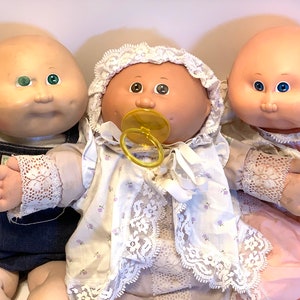 May include: Three Cabbage Patch Kids dolls, one wearing a blue overall, one wearing a white dress with a yellow pacifier, and one wearing a pink dress. All dolls have bald heads and large eyes.
