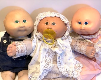May include: Three Cabbage Patch Kids dolls, one wearing a blue overall, one wearing a white dress with a yellow pacifier, and one wearing a pink dress. All dolls have bald heads and large eyes.