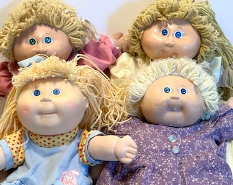 May include: Four Cabbage Patch Kids dolls with blonde yarn hair and blue eyes. The dolls are wearing different outfits, including a pink dress, a yellow dress, a blue dress with red polka dots, and a purple dress with white flowers.