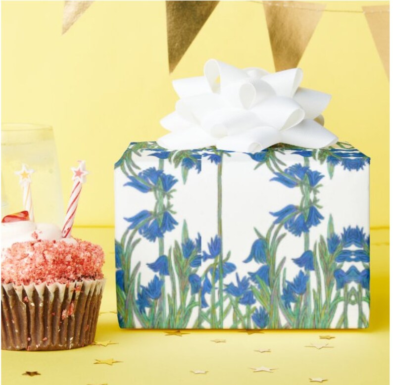 Shown above: A medium sized square gift box with the wrapping, and a white bow on top.  The box is sitting on a yellow surface with gold stars scattered around it and a cupcake with a candle sitting next to it.