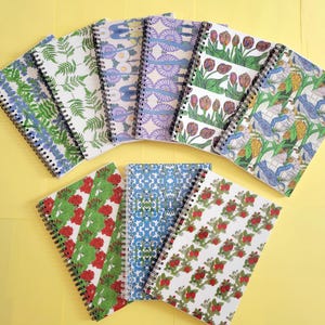 May include: A collection of eight spiral-bound notebooks arranged in a fan shape. Each notebook features a distinct cover design, including patterns of green ferns, purple floral motifs, red tulips, blue birds, red flowers, and red strawberries.