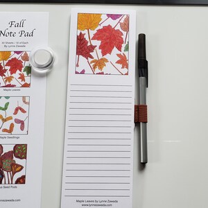 May include: A white notepad with lined pages and a colorful maple leaf design at the top. A pen with a brown band is next to the notepad. A smaller notepad with three images of fall leaves is held by magnets.