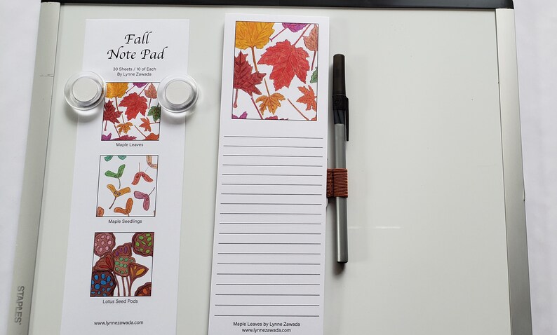 May include: A white notepad with lined pages and a colorful maple leaf design at the top. A pen with a brown band is next to the notepad. A smaller notepad with three images of fall leaves is held by magnets.