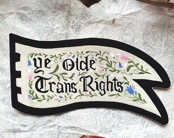 May include: A flag-shaped sticker with the text "ye Olde Trans Rights" in a vintage font, surrounded by floral designs in green, pink, and blue. The sticker has a black border and a cream-colored background.