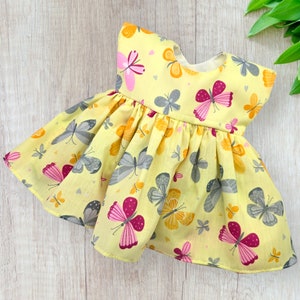 May include: A yellow dress adorned with a butterfly print. The dress has a gathered skirt and short sleeves. The butterflies are in pink, grey, and orange hues. The dress is made from a light fabric.