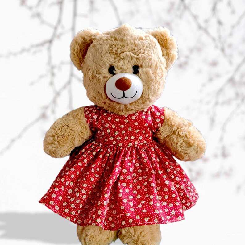 May include: A light brown teddy bear wearing a red dress with a white floral pattern. The dress has a gathered skirt and a sweetheart neckline.