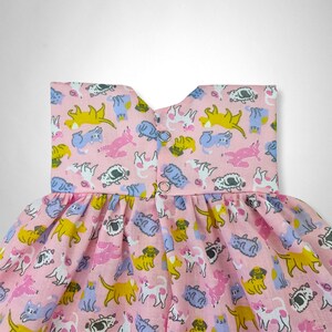 May include: A pink sleeveless dress with a ruffled skirt. The dress is covered in a pattern of cartoon cats in various colors, including yellow, blue, pink, and gray.