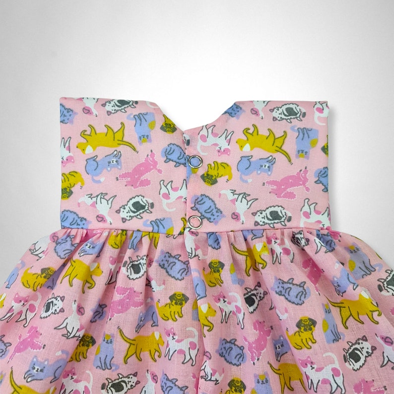 May include: A pink sleeveless dress with a ruffled skirt. The dress is covered in a pattern of cartoon cats in various colors, including yellow, blue, pink, and gray.