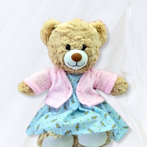 May include: A light brown teddy bear wearing a pink knitted cardigan and a blue dress with a floral pattern and bee designs.