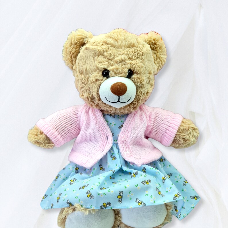 May include: A light brown teddy bear wearing a pink knitted cardigan and a blue dress with a floral pattern and bee designs.