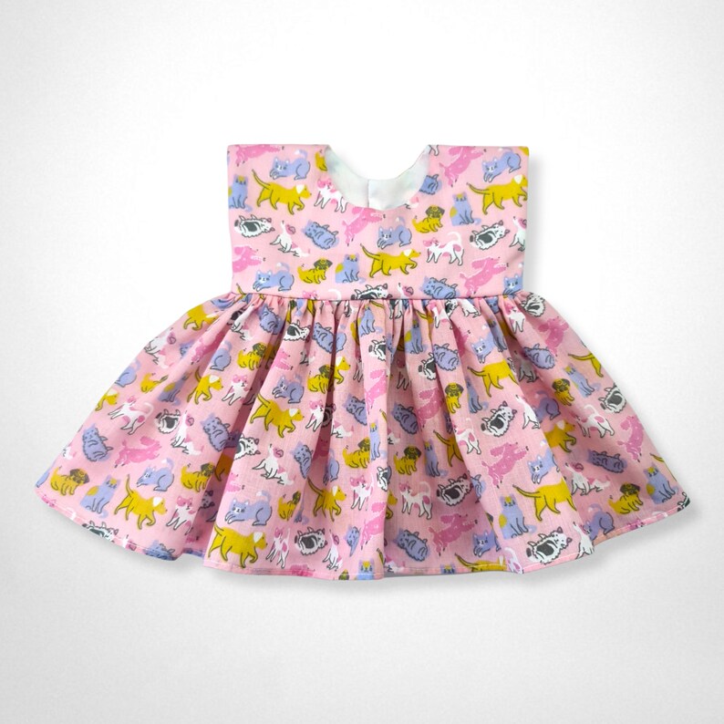 May include: A pink sleeveless dress with a gathered skirt. The dress is covered in a repeating pattern of colorful cartoon dogs and cats.