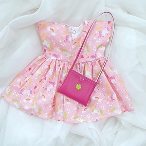 May include: A pink dress with a unicorn print and a matching pink purse. The dress has a white trim and a white bow at the neckline. The purse has a green flower on the front.