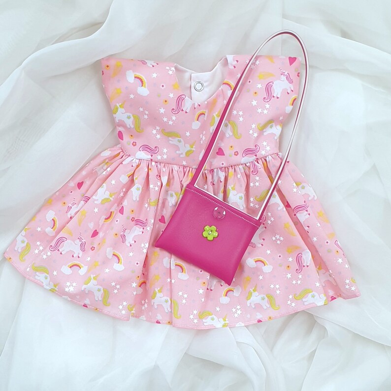 May include: A pink dress with a unicorn print and a matching pink purse. The dress has a white trim and a white bow at the neckline. The purse has a green flower on the front.