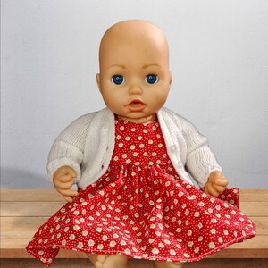 May include: A baby doll wearing a white cardigan over a red dress with a white floral pattern.