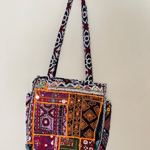 May include: A colorful patchwork tote bag with long handles. The bag features a variety of patterned fabrics in shades of purple, orange, red, and black, with decorative mirror accents. The handles have a repeating geometric design.
