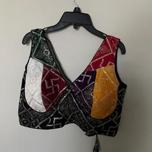 May include: A patchwork crop top with a V-neck design. The top features panels of various colors, including green, red, yellow, and purple, with intricate silver embellishments. The base fabric is black with a geometric pattern.