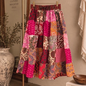 May include: A midi-length skirt featuring a patchwork design with various floral, paisley, and geometric patterns in shades of hot pink, deep red, mustard yellow, and cream. The skirt has an elasticized waistband with a drawstring tie.