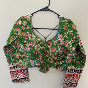 May include: Green long-sleeved blouse with intricate floral and peacock embroidery. The blouse features a V-neck and decorative trim on the cuffs. The embroidery includes pink, orange, blue, and gold accents. The blouse is hanging on a black hanger.