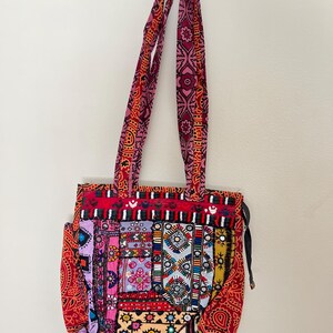 May include: A colorful patchwork tote bag with long straps. The bag features a vibrant mix of red, pink, purple, and yellow fabrics with intricate patterns and embellishments. The straps are a patterned red and pink fabric.