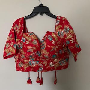 May include: A red, embroidered top with a deep V-neck and short sleeves. The top is adorned with gold, teal, and silver floral patterns. It has decorative tassels at the hem and is hanging on a black hanger.