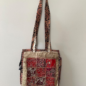 May include: A patchwork tote bag with a patterned strap. The bag features a square design with red, burgundy, and gold fabric squares. The strap is black with an orange and white paisley design. The bag has a gold trim.