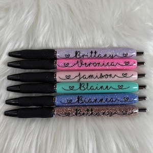 May include: Six glitter pens with black grips and silver accents. Each pen is a different color: purple, pink, teal, blue, and brown. Each pen has a name written on it in cursive with a heart symbol.