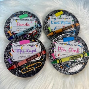 May include: Four round coasters with a composition book design. Each coaster features a name, a crayon, and a paperclip. The coasters are black with white speckles and a white border. The crayons are different colors, including blue, orange, pink, and yellow.