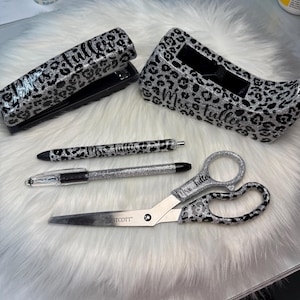 May include: A desk set featuring a stapler, pen holder, two pens, and scissors. The items have a silver glitter leopard print design with the name "Mrs. Fuller" on them. The scissors are branded "STCOTT". The items are on a white fluffy surface.