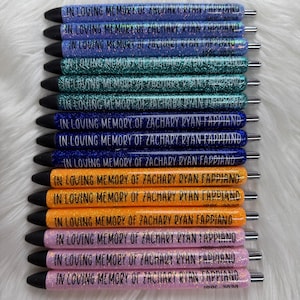 May include: A collection of glitter pens in various colors, including blue, green, orange, and pink. Each pen is inscribed with the text "IN LOVING MEMORY OF ZACHARY RYAN FAPPIANO 1995-2020". The pens have black tips and silver accents.