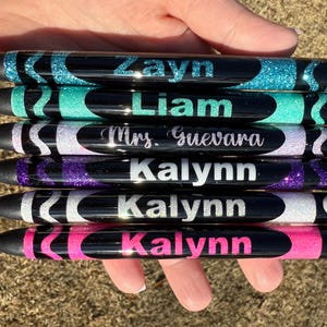 May include: Six black and glitter pens with names written on them. The names are Zayn, Liam, Mrs. Guevara, Kalynn, Kalynn, and Kalynn.
