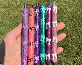 May include: Six glitter pens in various colors, including purple, coral, green, and pink. Each pen features a bow design. The pens are held in a hand against a background of green grass.