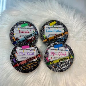 May include: Four round coasters with a composition book design. Each coaster features a name, a Crayola crayon, and a paperclip. The coasters are black with colorful accents and are set on a fluffy white surface.