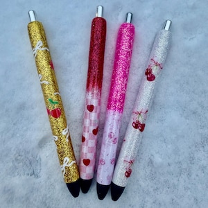 May include: Four glitter pens with different designs. The pens are gold, red, pink, and white with glitter. The designs include cherries, bows, and hearts.