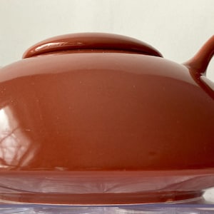 May include: A terracotta-colored ceramic teapot with a rounded body and a wide, flat spout. The teapot has a lid and a single handle.