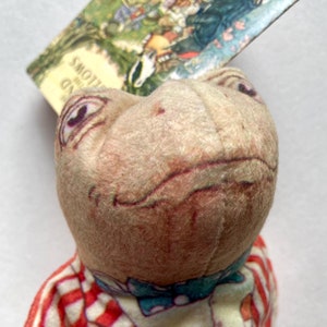 May include: A plush toy frog wearing a red and white striped shirt, a blue bow tie, and blue jeans. The frog has a brown and white face with a slight smile. The frog is holding a book titled "The Wind in the Willows" by Kenneth Grahame.
