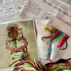 May include: An assortment of embroidery supplies, including a chart, instructions, and a design featuring a mouse in a dress. The kit includes various colored threads, a needle, and a color chart. The text on the chart reads "CREWEL EMBROIDERY STITCH CHART".