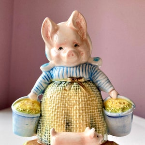 Puede incluir: Una alcancía musical de cerámica en forma de cerdo con un vestido a rayas azules y blancas, sosteniendo dos cubos de flores amarillas. Un pequeño cerdo rosa está de pie en la base de la alcancía, que está decorada con hierba verde.