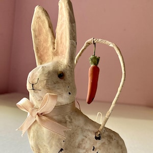 May include: A vintage-style Easter bunny figurine with a cream-colored finish and a pink ribbon bow. The bunny holds a carrot suspended from a curved wire. The figurine has black spots and is a charming Easter decoration.