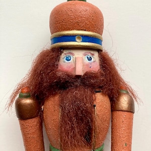 Könnte beinhalten: Eine handgefertigte Nussknacker-Puppe aus Holz mit einem strukturierten orangefarbenen Körper, einem roten Bart und einer detaillierten Krone. Die Puppe hat blaue Augen, eine rosa Nase und einen grünen und goldenen Gürtel. Der untere Teil der Puppe ist kariert.