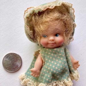 May include: A vintage doll with blonde hair and blue eyes, wearing a mint green dress with a polka dot pattern and lace trim. The doll is wearing a matching bonnet. A quarter is next to the doll for size comparison.